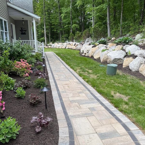Stone walkway beside a house with garden and trees.