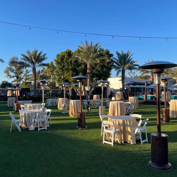 Outdoor event setup with tables, chairs, heaters, and palm trees under clear blue sky.