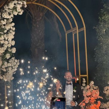 Newlywed couple smiling under a floral arch with sparklers at night.
