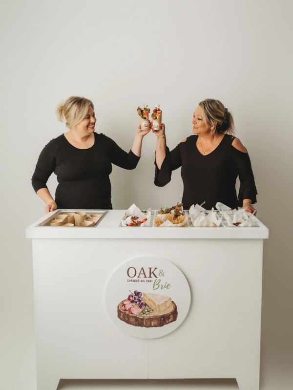 Two women cheers with appetizers behind a charcuterie stand.