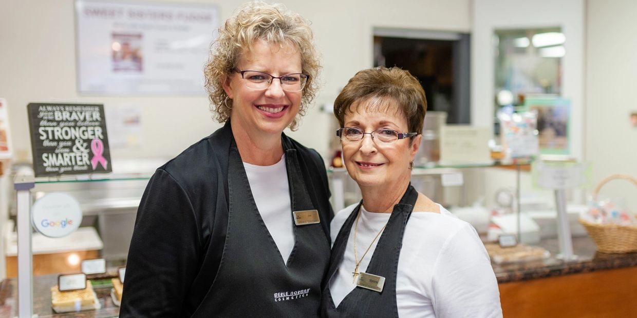 Two smiling women in aprons standing in a bakery or café.