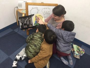 Woman reading a picture book to three children sitting on the floor.