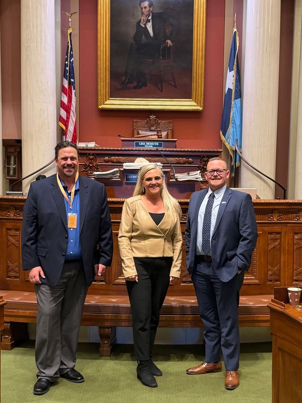 Three professionals pose in a formal government chamber with an Abraham Lincoln portrait.