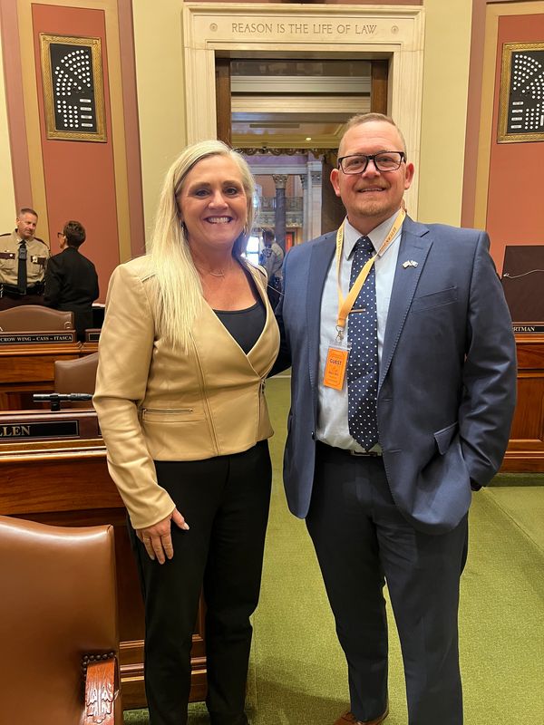 Two people smiling inside a formal legislative chamber with 'Reason is the Life of Law' inscribed above.