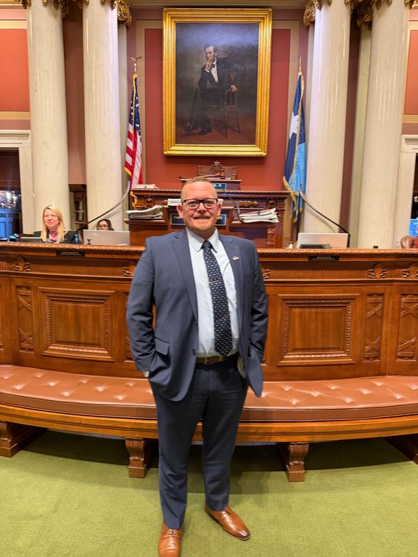 Man in a suit standing in a formal government chamber with a painting behind.