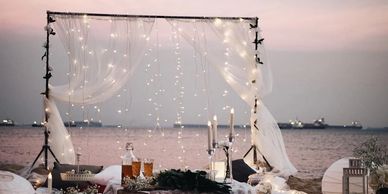 Romantic beachside dinner setup with fairy lights and candles at sunset.