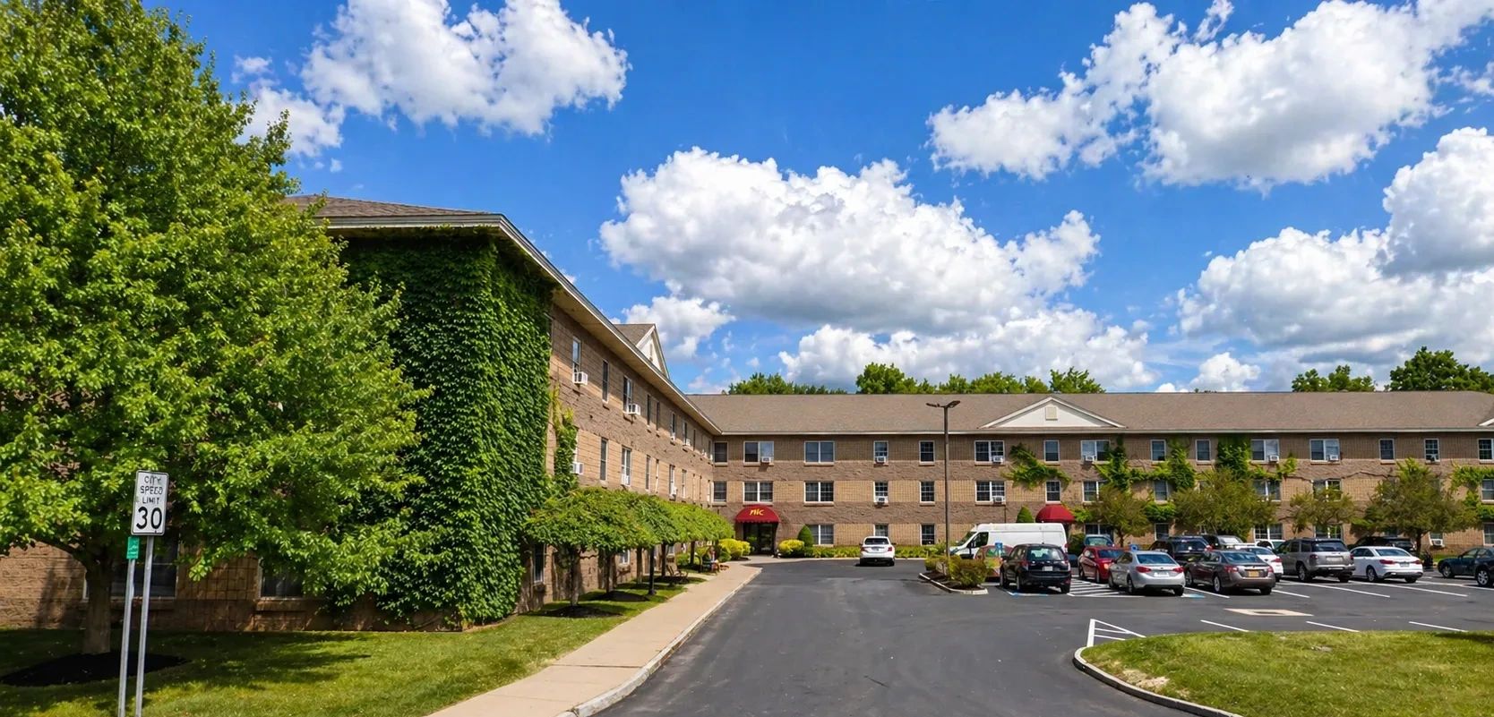 A large brick building with greenery and a parking lot under a bright blue sky.