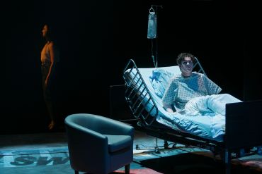 Patient in hospital bed with a standing woman in the shadows.