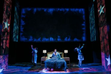 A person sits on a bed with two others dancing nearby on a dramatic stage.