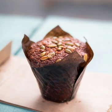 A close-up of a pumpkin seed-topped muffin in a brown parchment wrapper.