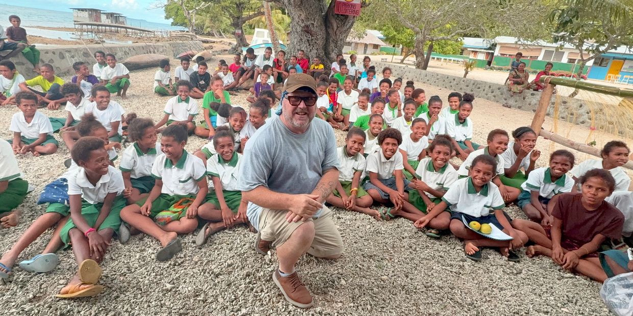 Man kneeling with a large group of smiling schoolchildren outdoors by the sea.