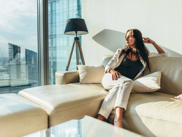 A woman in a white suit sits on a light-colored sofa, looking out a large window at a city skyline.