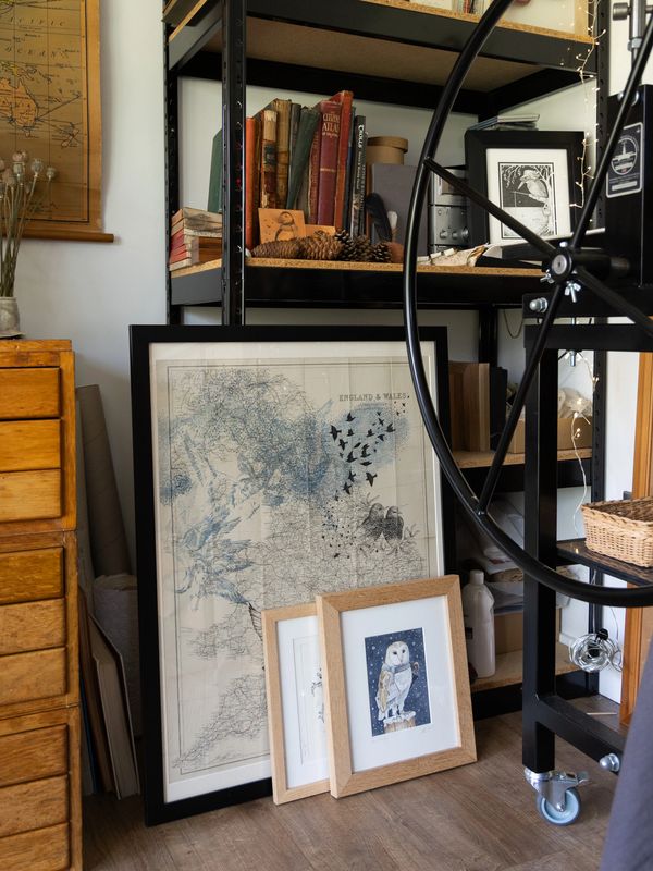 Art-filled cozy corner with framed maps, owl prints, and vintage books on shelves.