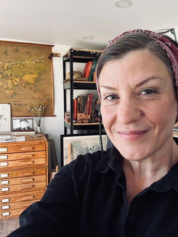 A woman smiling in a cozy room with maps and books in the background.