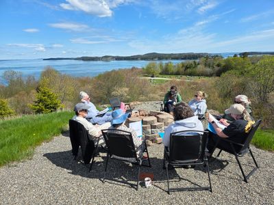 Group of people sitting around a fire pit overlooking a scenic waterfront.