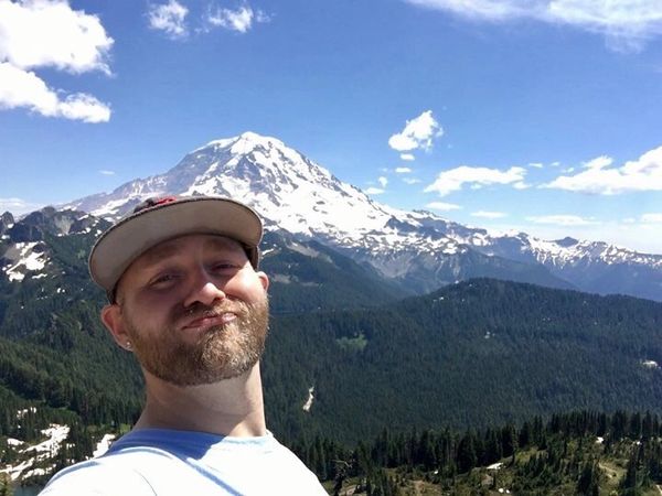 Man taking selfie with snow-capped mountain and forest background.