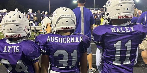 Three young football players in purple jerseys on the sidelines.
