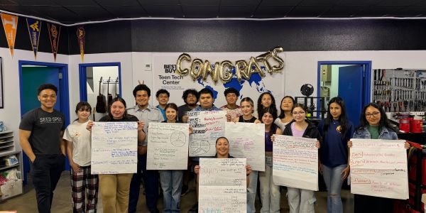 Group of smiling students holding charts beneath CONGRATS balloons.
