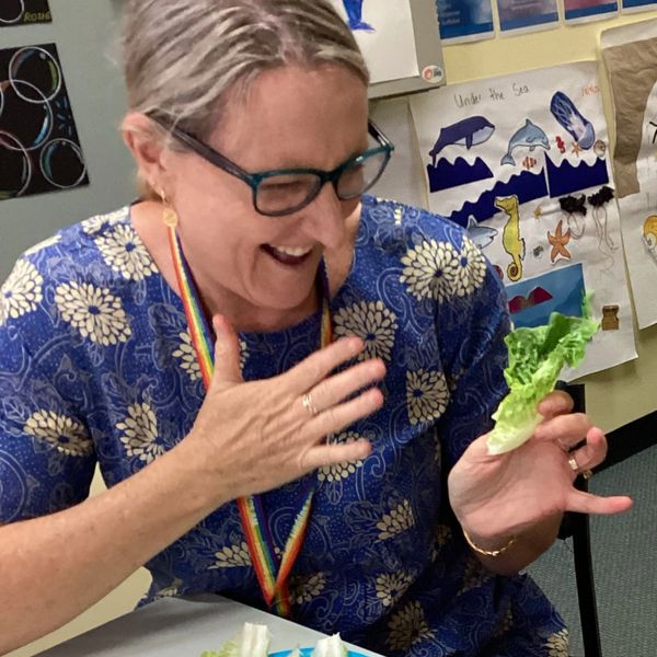Karin in a blue dress with white flowers, wearing blue glasses is signing the Auslan sign for "like"