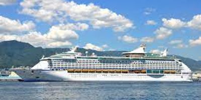 A large cruise ship sailing near mountainous coastline under a blue sky.