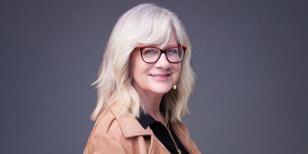Smiling woman with white hair and red glasses wearing a brown jacket.