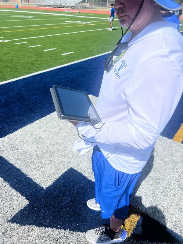 Coach in white hoodie and blue shorts holding a tablet on a football field.