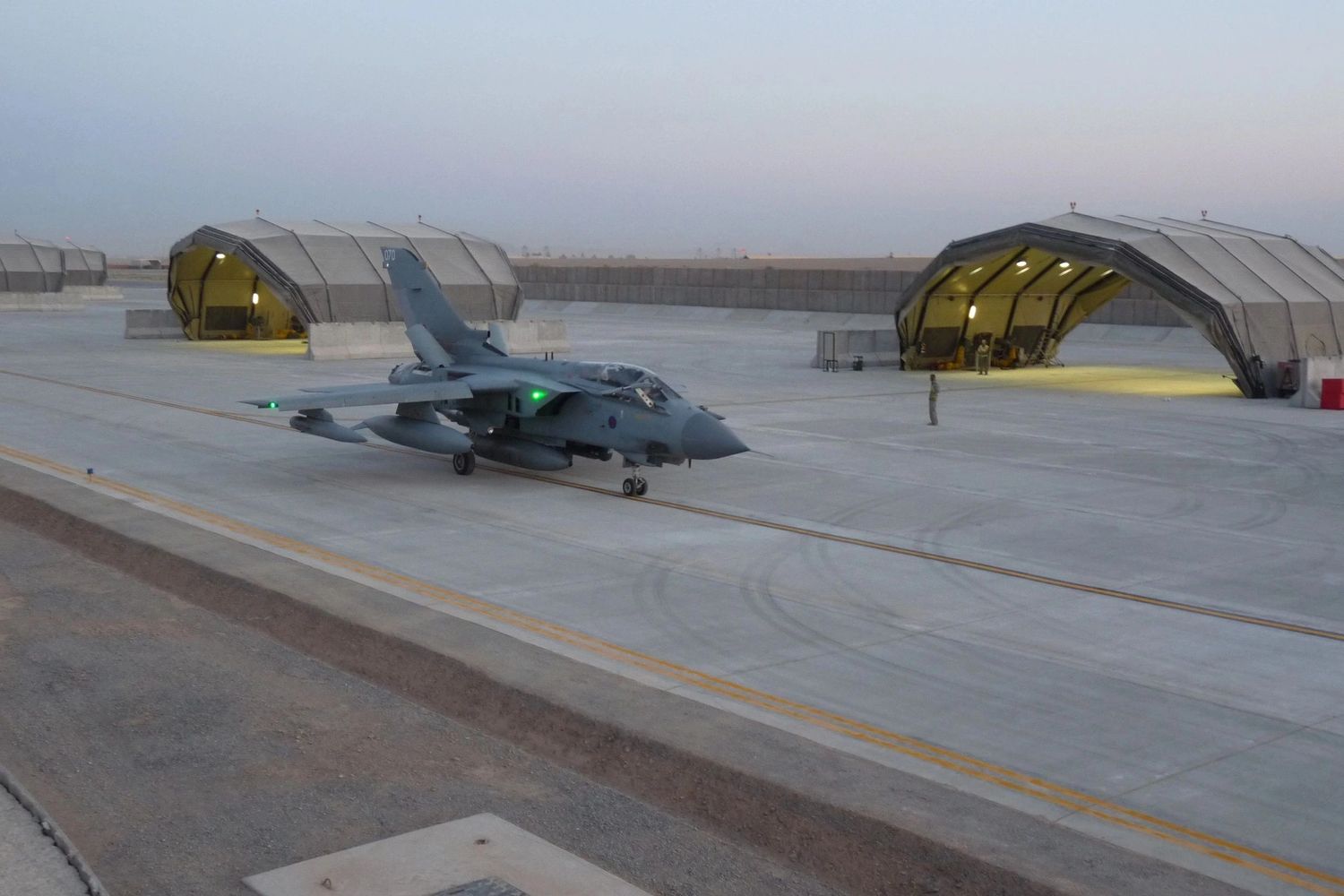 Military jet taxiing on concrete runway near shelters at dusk.