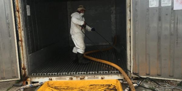 Worker in protective gear cleaning inside a metal container with hose and water.