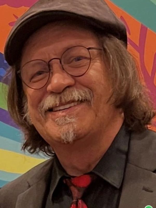 Smiling man with glasses, a cap, and a colorful background.