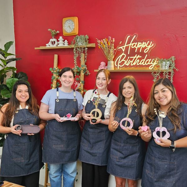 Five women in aprons holding crafts under a Happy Birthday sign.