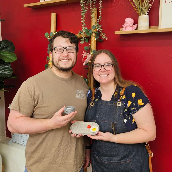 A smiling couple holding handmade pottery in a cozy, decorated room.