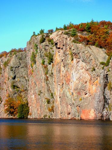 Tall rocky cliff with autumn trees above calm water under a clear blue sky.