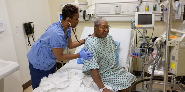 Elder man or woman in a hospital bed being helped by a nurse.