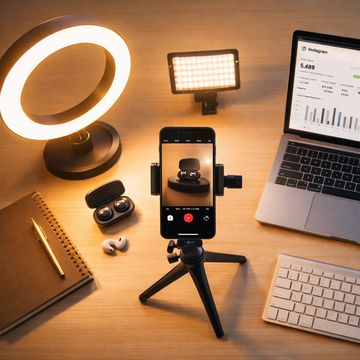 A smartphone on tripod filming wireless earbuds with ring light and laptop on desk.