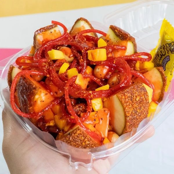 A hand holding a spicy fruit salad with chamoy and chili powder in a plastic container.