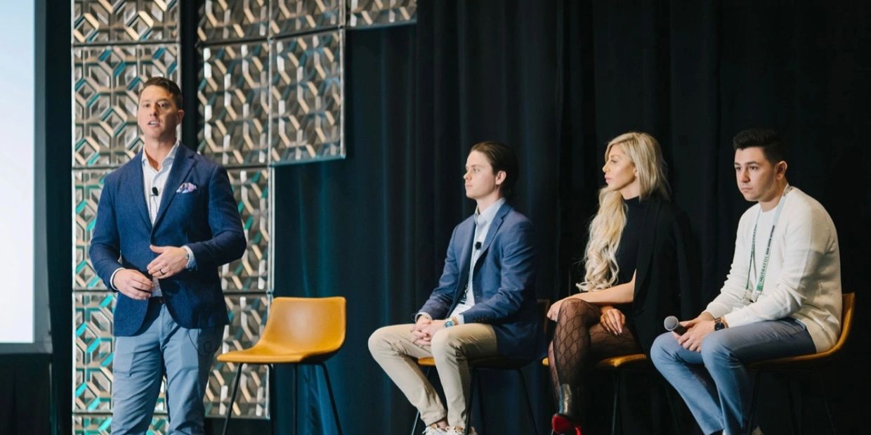 A man speaks while three people sit on stage at a conference or panel discussion.