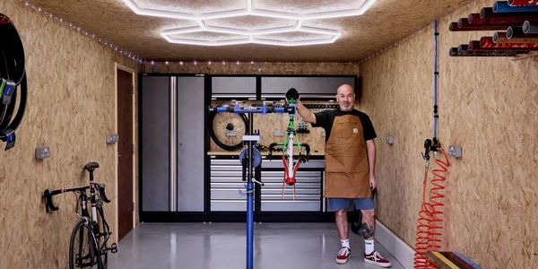 Man in a workshop holding a bike frame, surrounded by tools and bikes.
