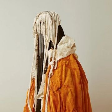 Abstract figure draped in layered, tattered fabrics with earthy tones.