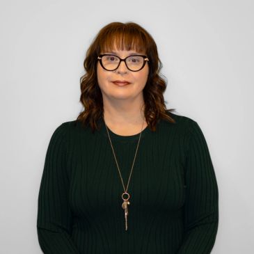 Woman with glasses and necklace against plain background.
