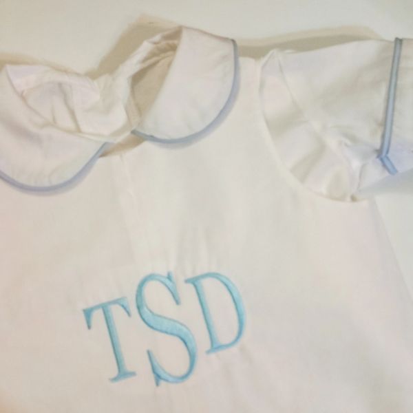 White garment with blue embroidered initials TSD and a ruffled collar.