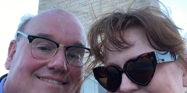 A smiling man and woman wearing glasses taking a selfie outdoors.