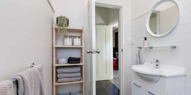 Compact bathroom with white walls, a round mirror, and wooden shelves with towels and decor.