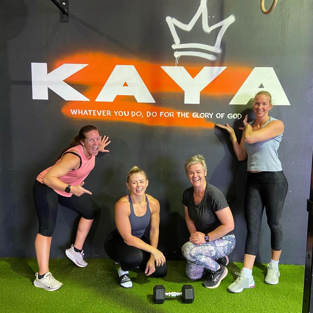 Four women posing in workout clothes by a motivational gym wall.