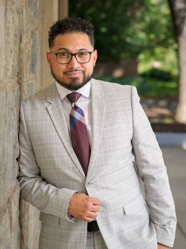 Man in a light gray plaid suit leaning against a stone wall outdoors.