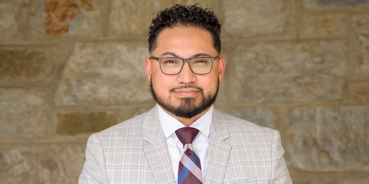 Man in a light gray checkered suit with a burgundy tie stands confidently against a stone wall.