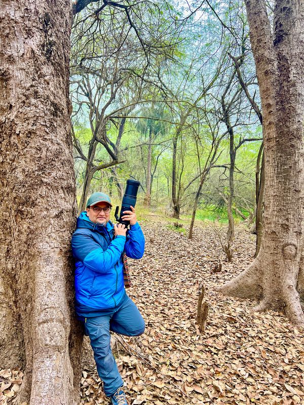 Man in blue jacket holding a camera in a forest with dry leaves.