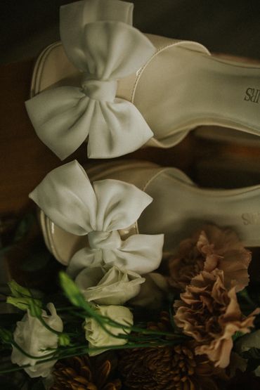 Elegant cream heels with large bows beside soft flowers.