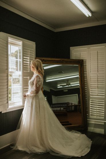 Bride in a lace wedding gown looks out a window in a softly lit room.