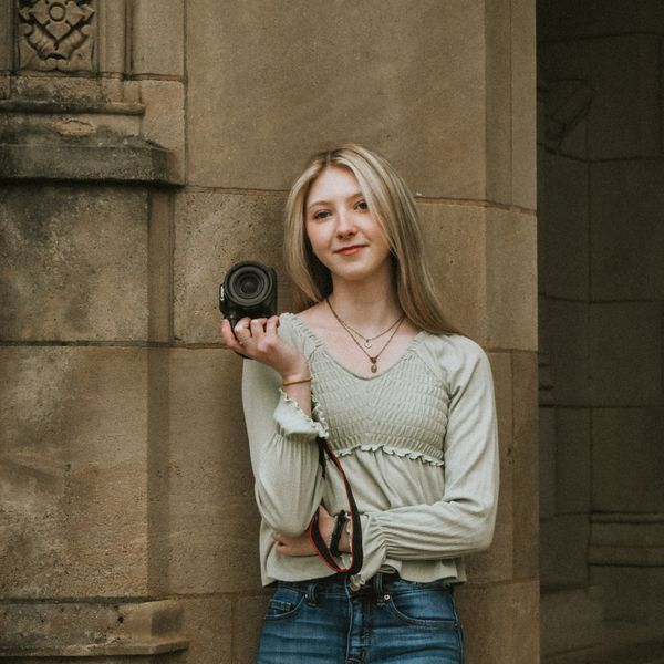 Young woman holding a camera, leaning against a stone wall.