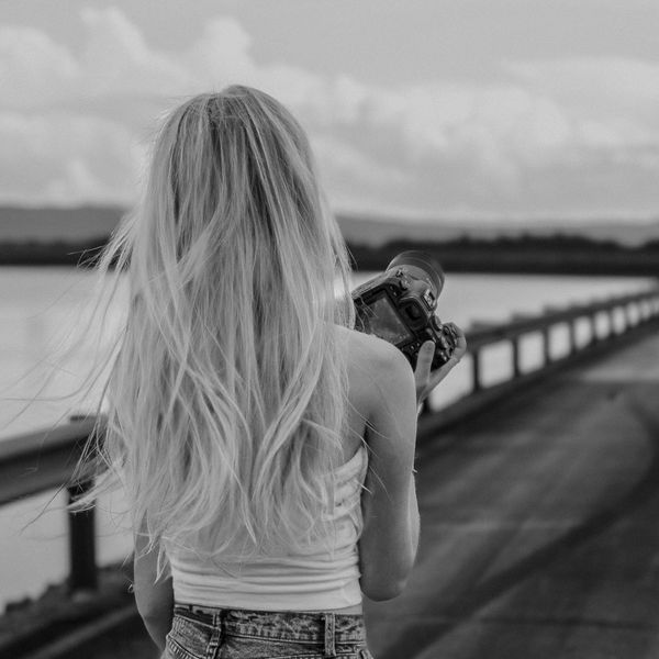 Woman with long hair holding a camera on a bridge in black and white.